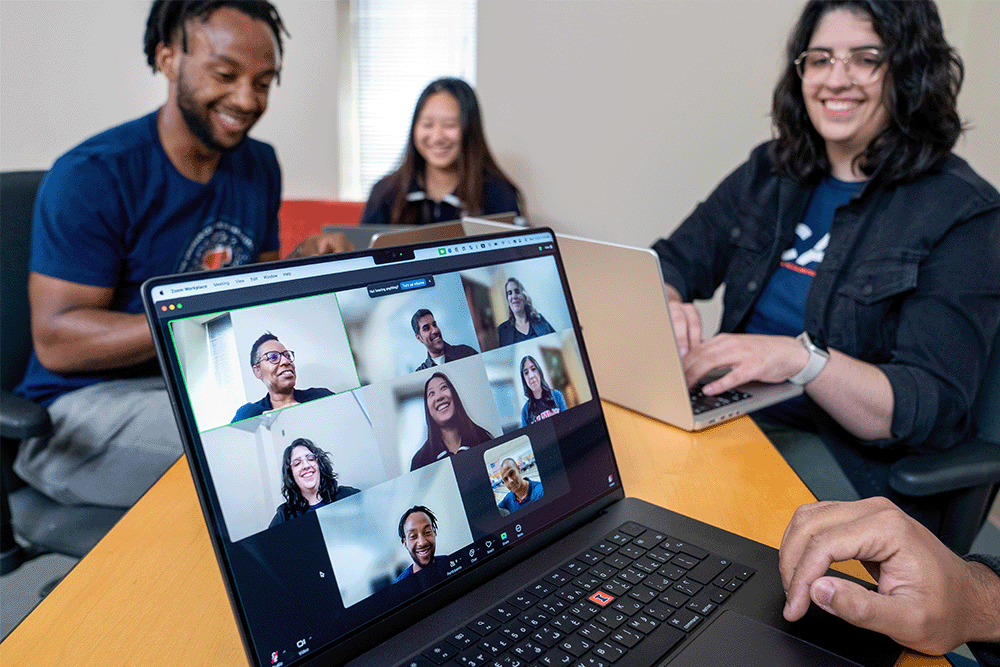 A laptop sits in the middle of the table, with small squares containing talking and smiling students, surrounded by three seated students and two seated adults talking and laughing with them.