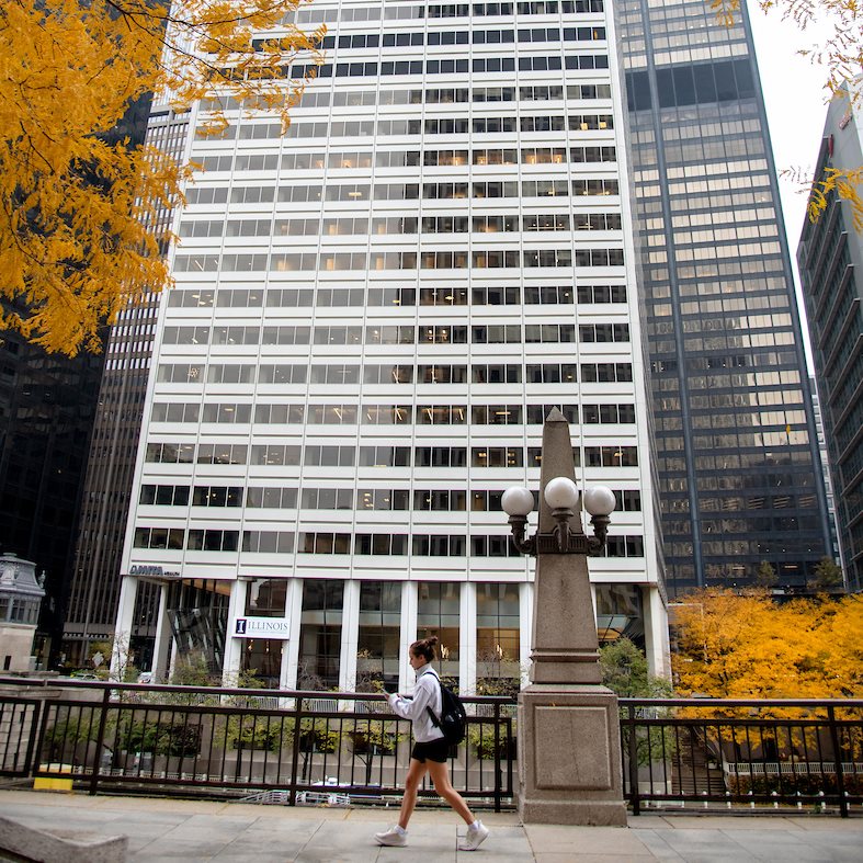 A white high-rise in a city with an orange tree and a person walking by a lamp post.