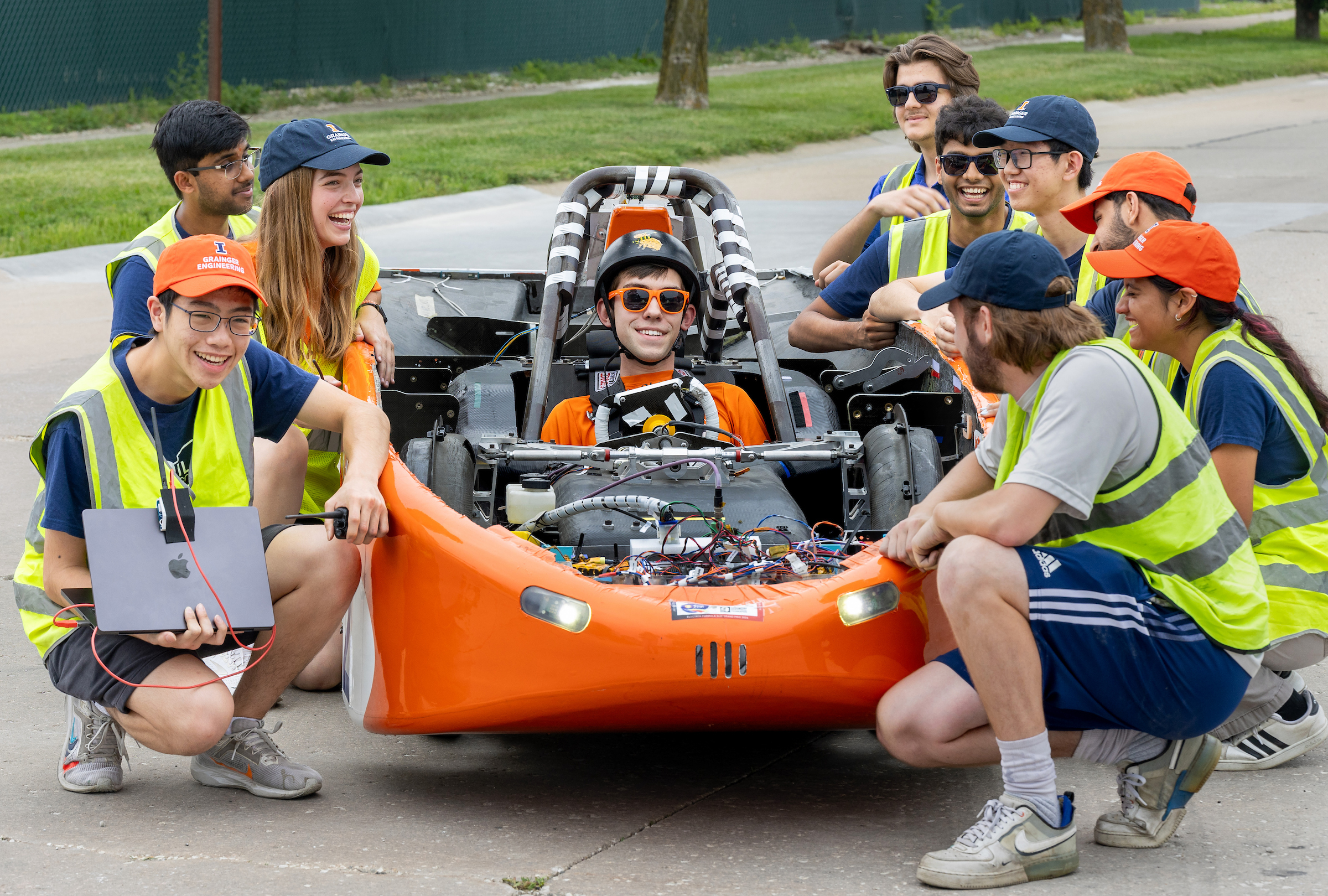 People smile and crouch around an orange solar car with a driver in a helmet, smiling.