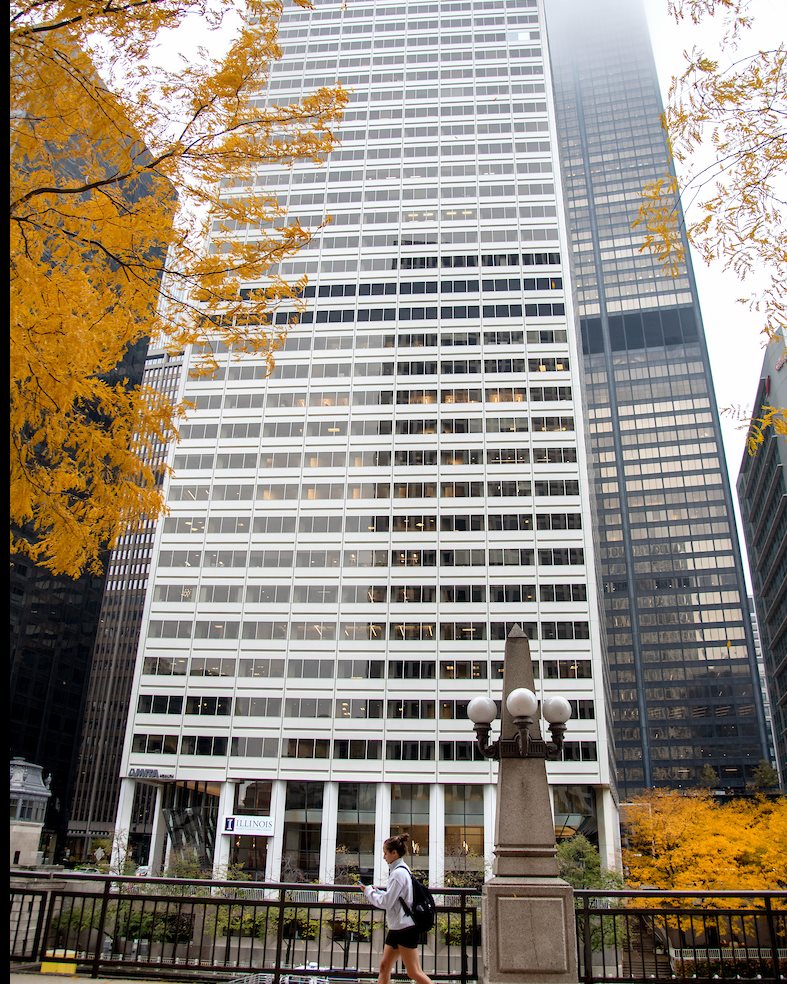 A white high-rise in a city with an orange tree and a person walking by a lamp post.