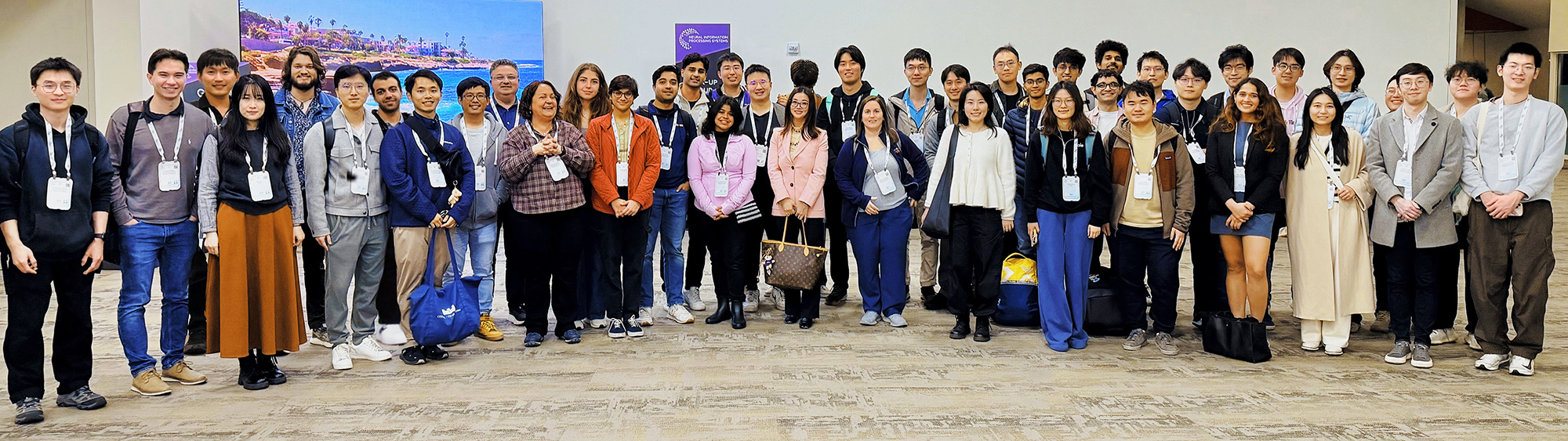 A large group of people stands against a white wall at a conference, smiling.