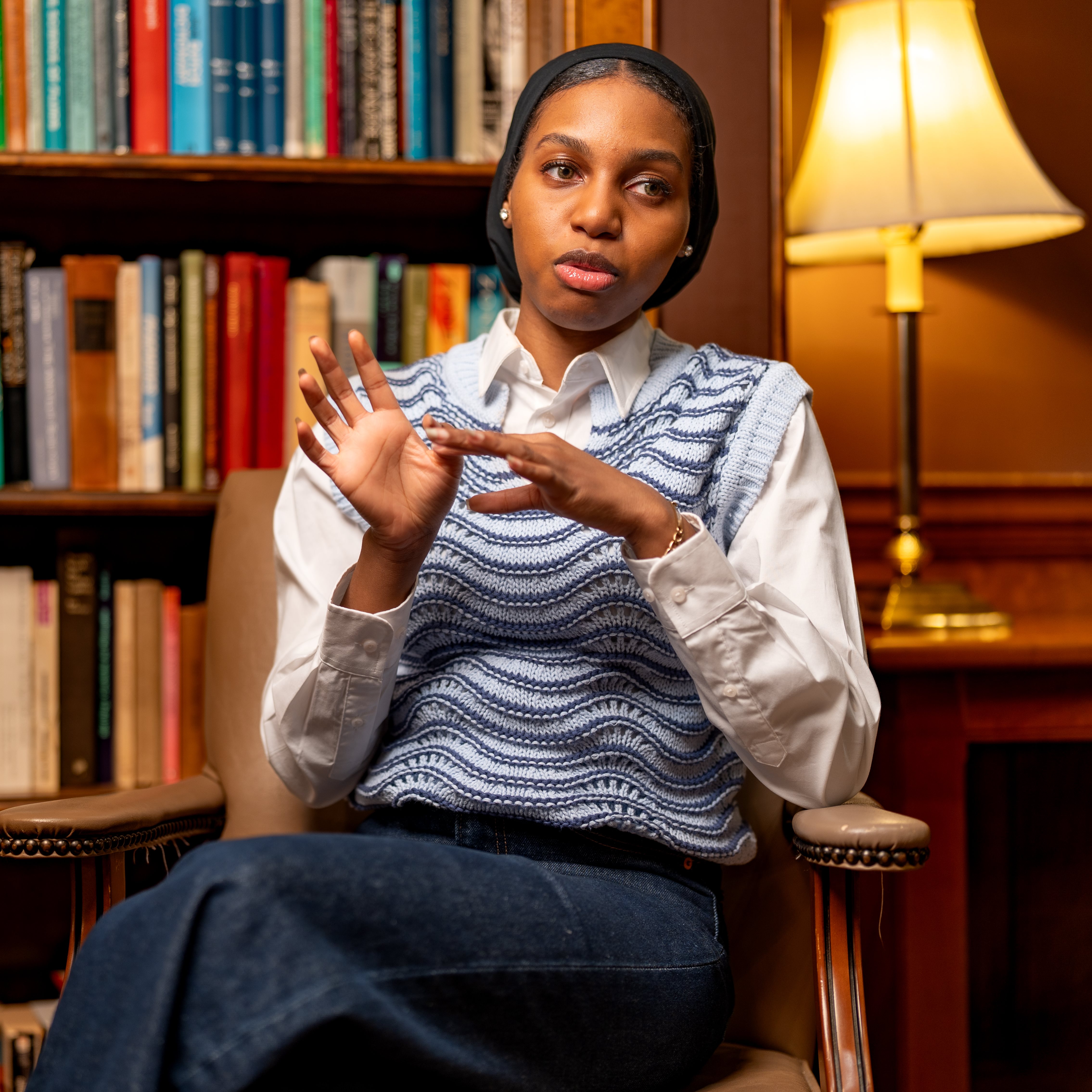 A woman sits in front of a stack of books and gestures.