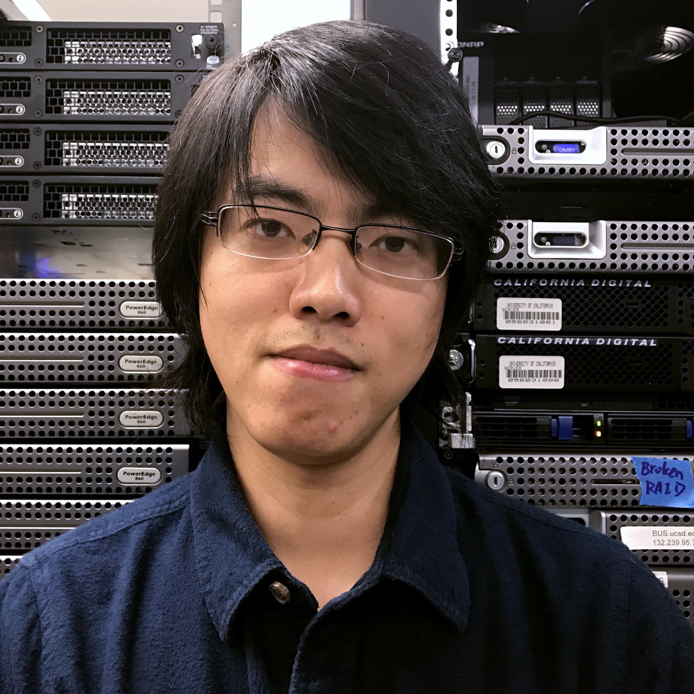 Headshot of professor Tianyin Xu, in a dark shirt with glasses standing in front of computer equipment.