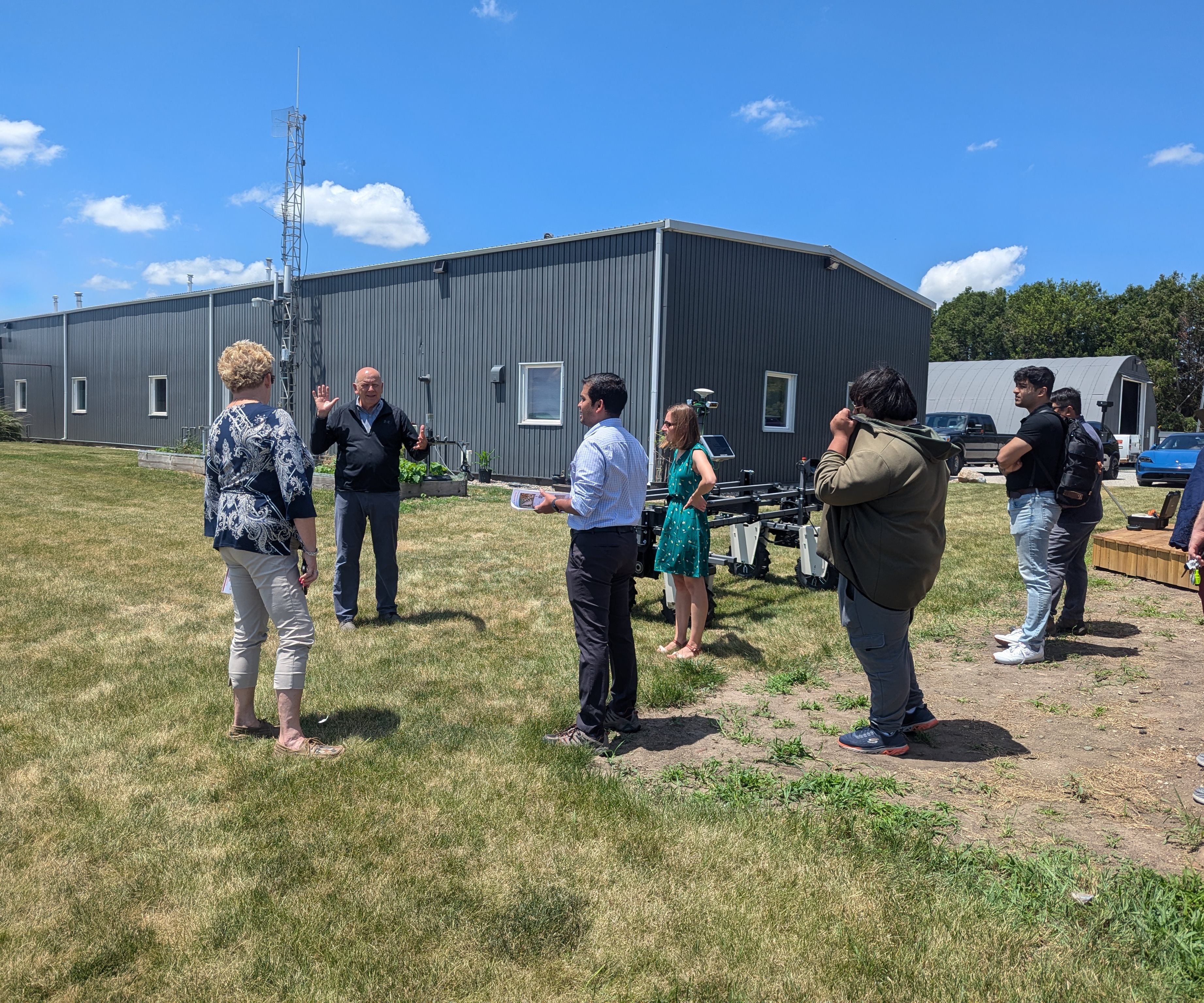 Several people stand in grass as John ried gives a presentation to the other people.