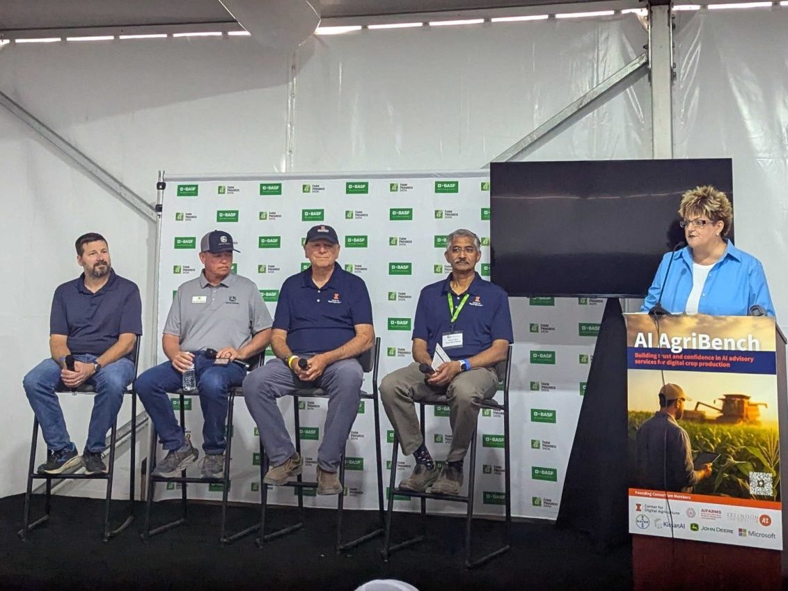 Five people are on a stage: four people are in chairs and one is behind a podium.