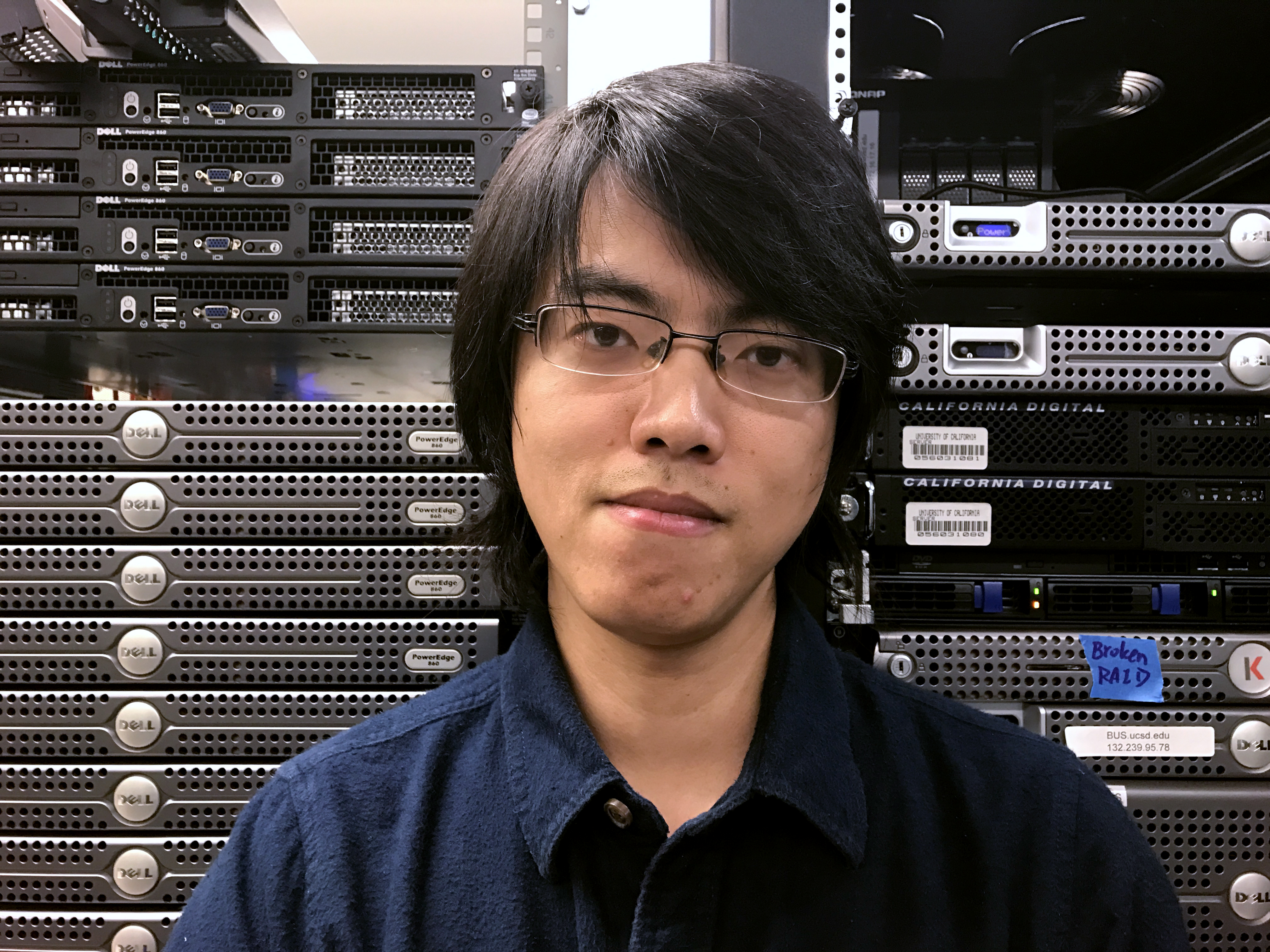 Headshot of professor Tianyin Xu, in a dark shirt with glasses standing in front of computer equipment.