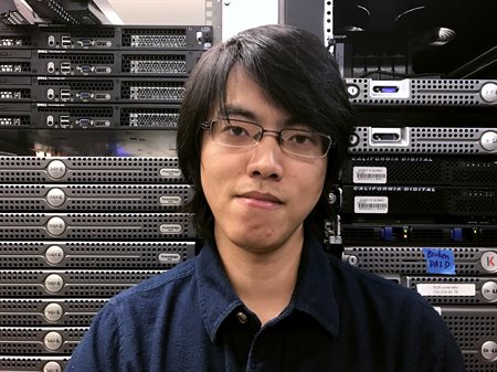 Headshot of professor Tianyin Xu, in a dark shirt with glasses standing in front of computer equipment.