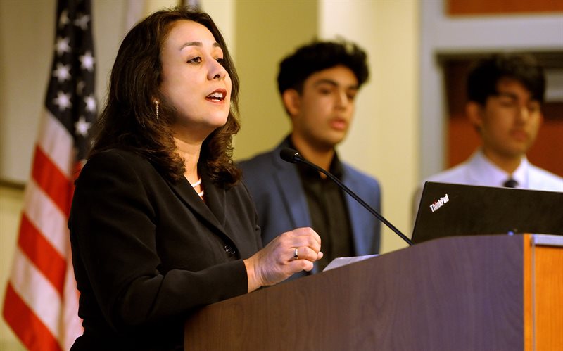 Sanchita Banerjee Saxena stands at a podium - with her two sons standing behind her, as well as an American flag - speaking at the Inaugural Banerjee Lecture.