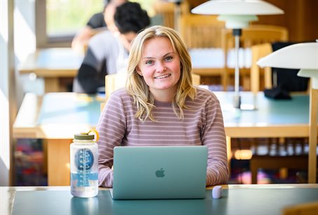 A woman sitting in a library with a water bottle looks up from her laptop.