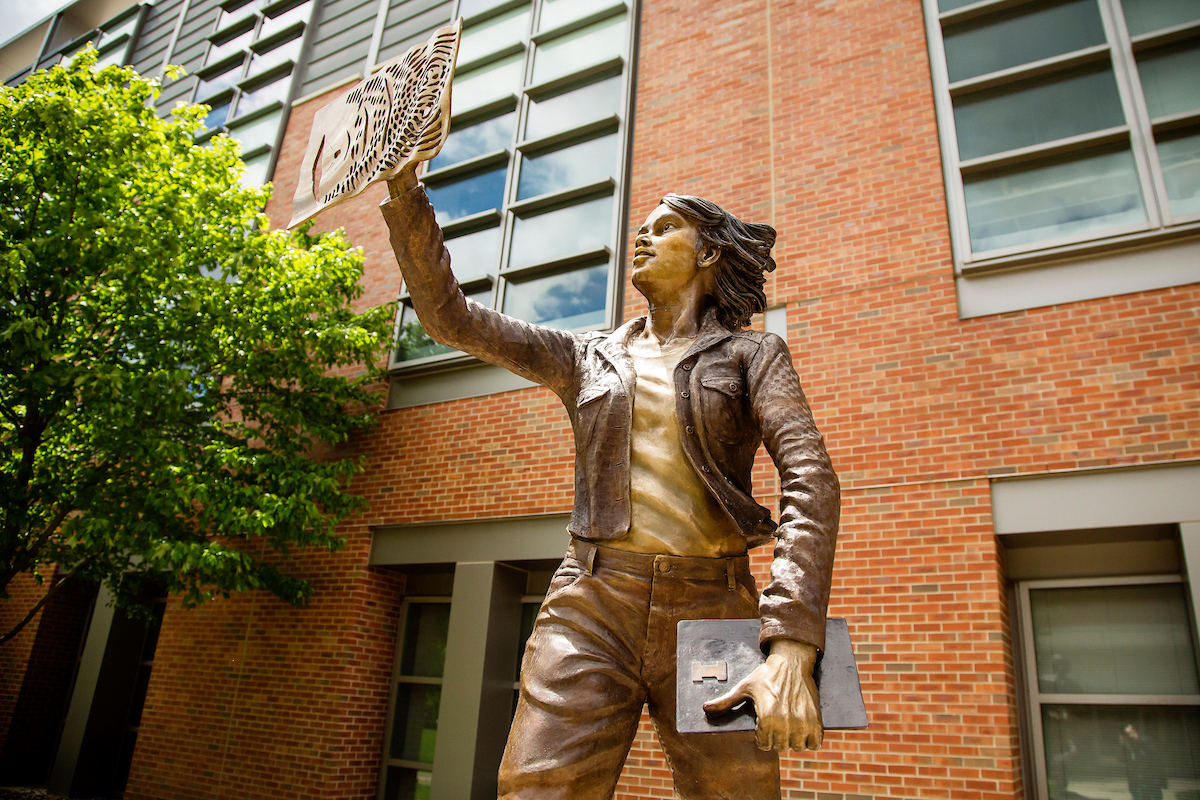 Bronze sculpture of a woman reaching upwards and touching a computer screen while holding a book or laptop with a large Block I on it.