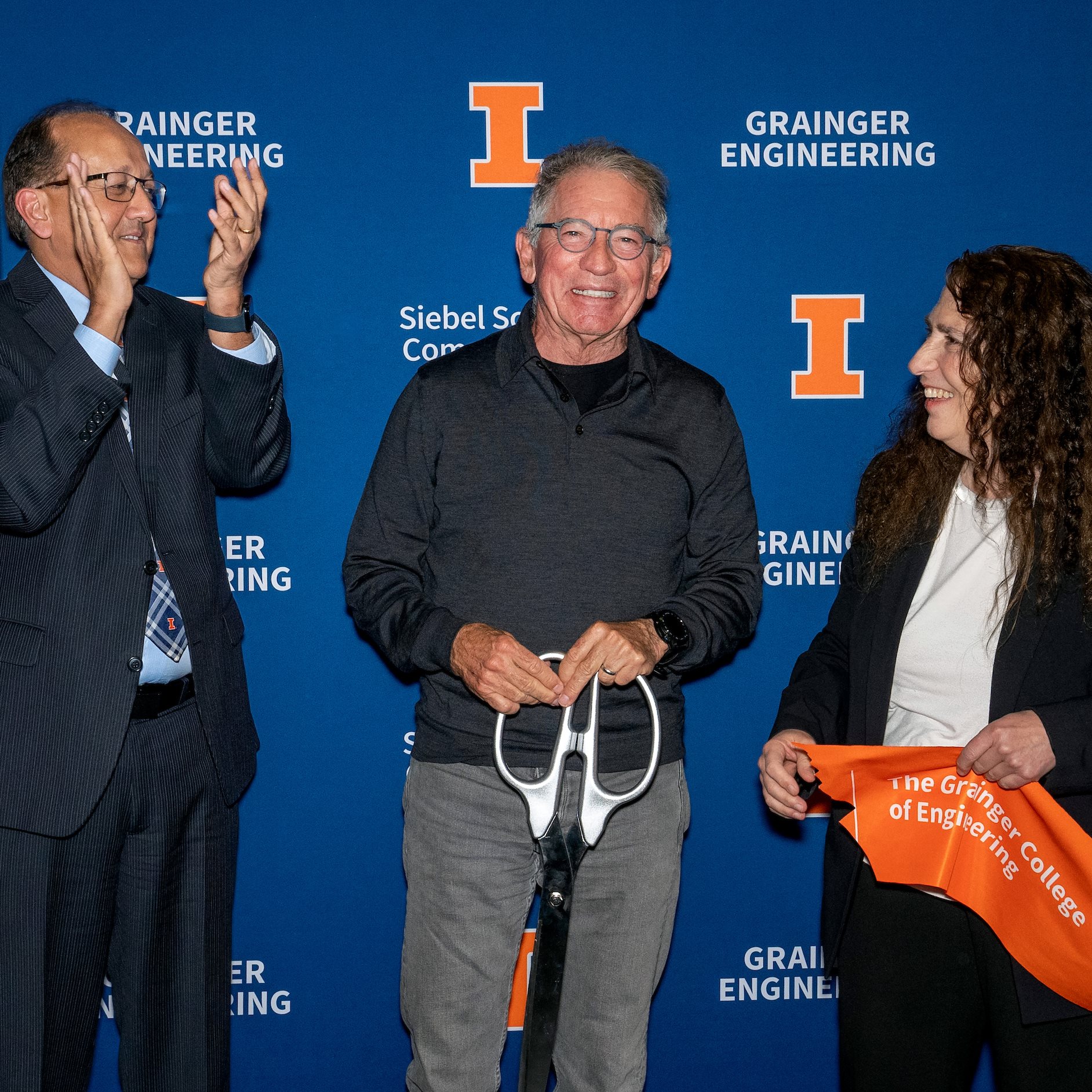 One person claps, the other smiles, and the other holds a giant pair of scissors in front of a blue background. 