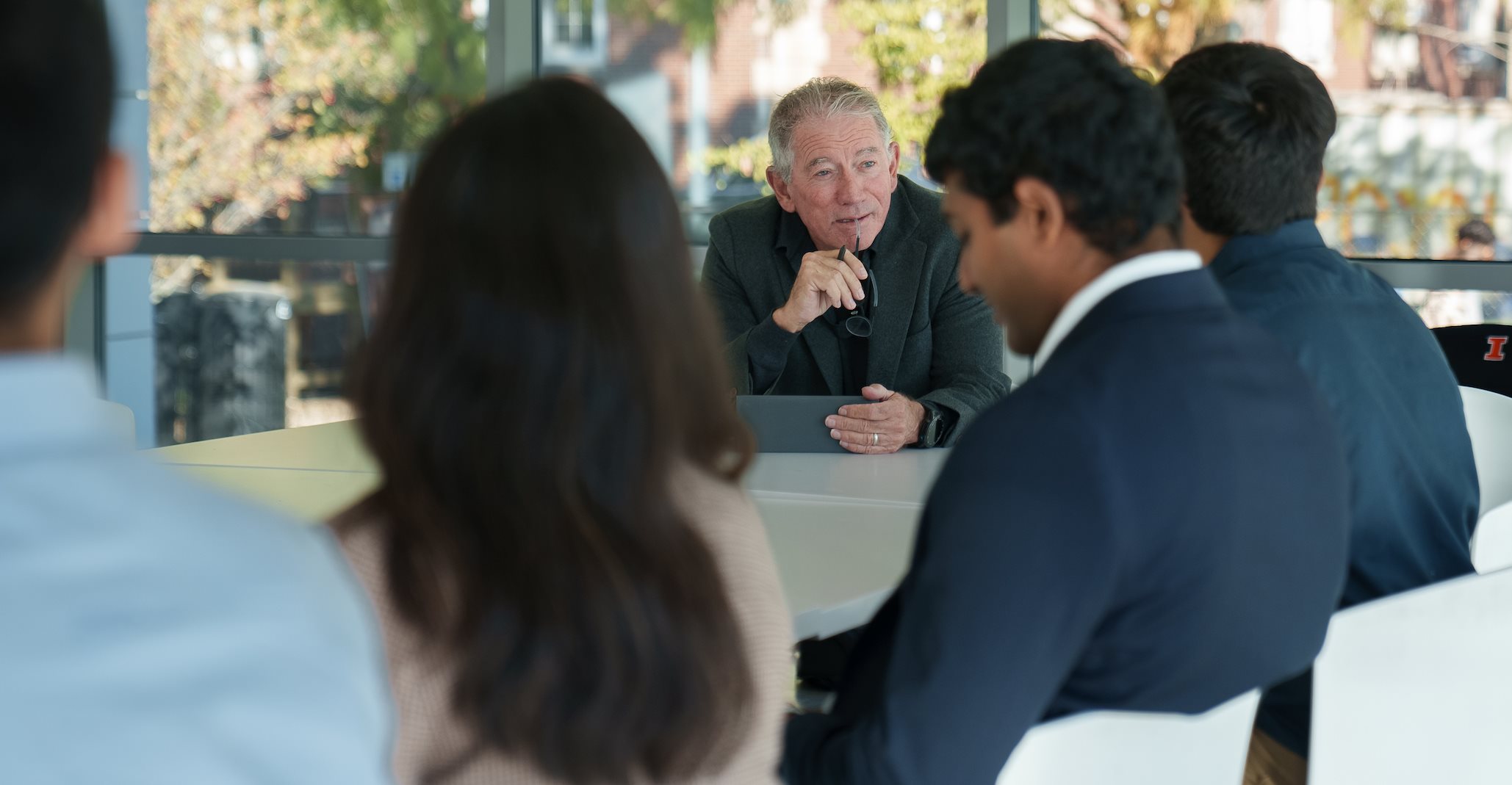 A man sits and talks to people in front of him. Green trees are in the background.