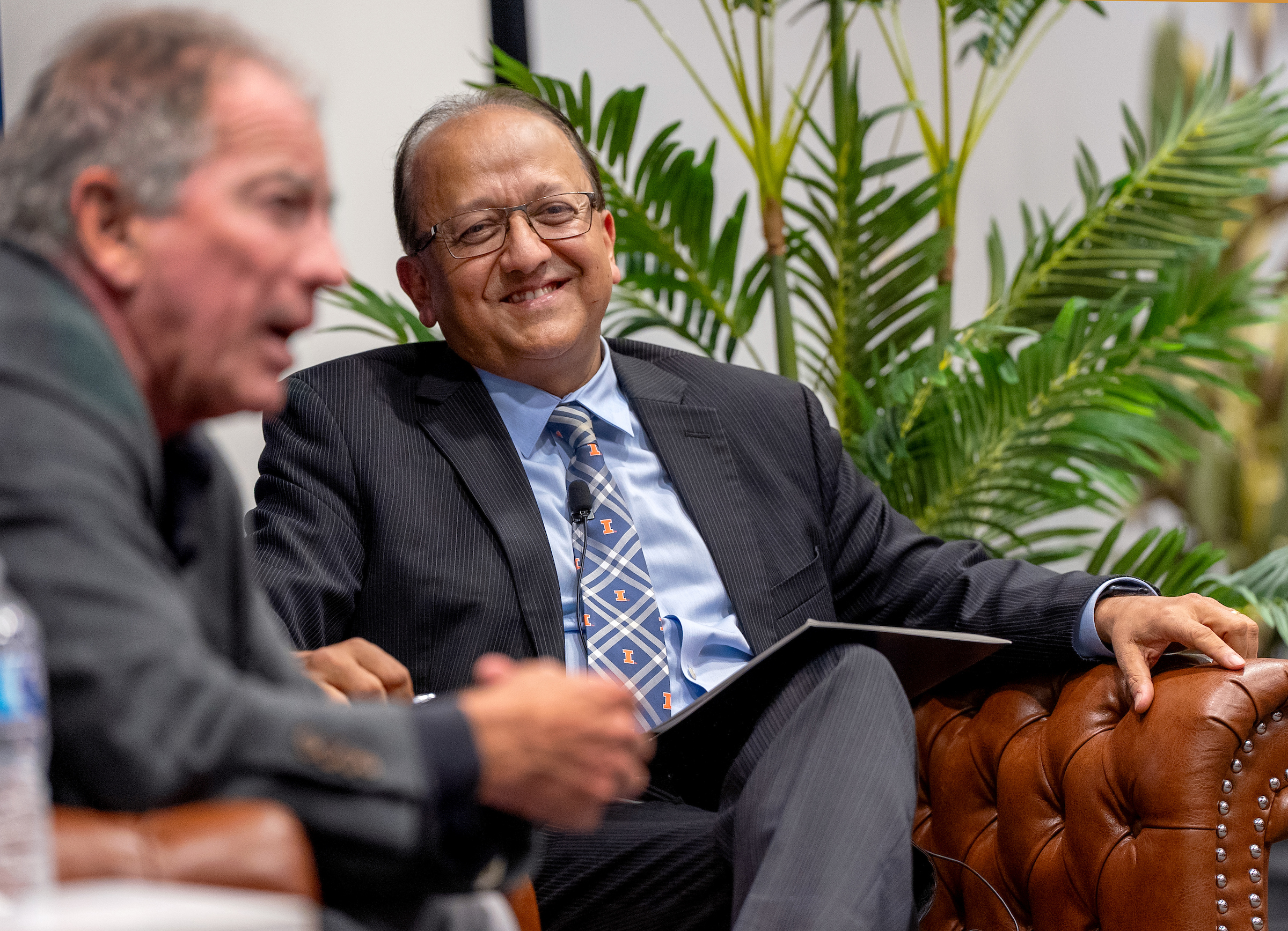 Two men sit and laugh in front of a large green plant. 